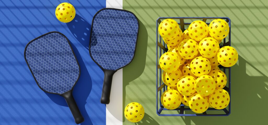 Two,Pickleball,Paddles,And,Basket,Of,Balls,On,Court,Under Rackets and balls on a pickleball court